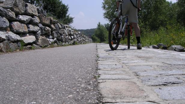 Ein Radfahrer mit Rucksack steht auf einem Weg aus Kopfsteinpflaster neben einer asphaltierten Strecke und einer Steinmauer.