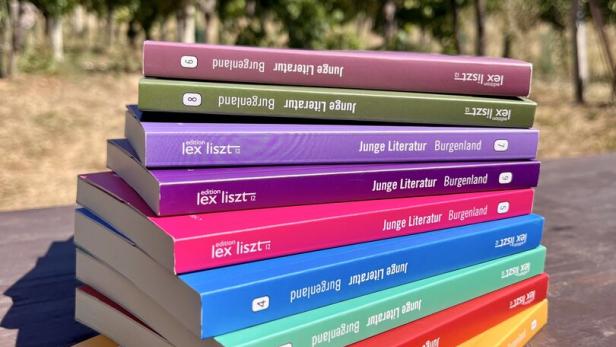 Ein Stapel bunter Bücher der Reihe „Junge Literatur Burgenland“ der edition lex liszt 12 liegt auf einem Holztisch vor einem Weinberg.