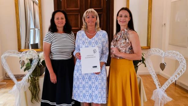 Drei Frauen (in der Mitte Stadtchefin Böhm) stehen nebeneinander, wobei die mittlere eine Urkunde der Stadt Neusiedl am See hält, flankiert von zwei weißen Herz-Dekorationen.