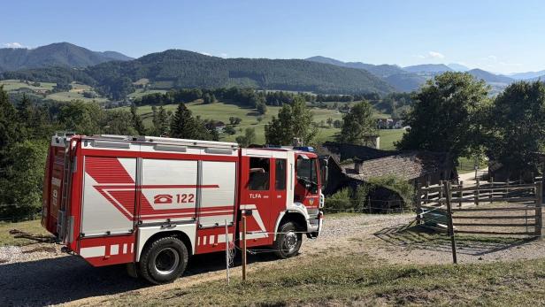Ein rotes Feuerwehrfahrzeug steht auf einem Feldweg vor einer ländlichen Bergkulisse.