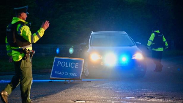 Polizisten sperren nachts eine Straße, ein Auto mit Blaulicht steht vor einem Schild „POLICE ROAD CLOSED“.