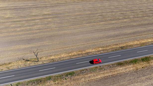 Ein rotes Auto fährt auf einer einsamen Landstraße durch eine weitläufige, trockene Ackerlandschaft.