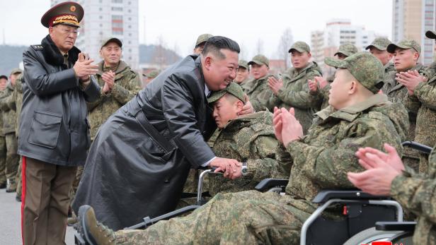 Kim Jong-un umarmt einen sitzenden Soldaten, umgeben von applaudierenden Militärangehörigen im Freien.