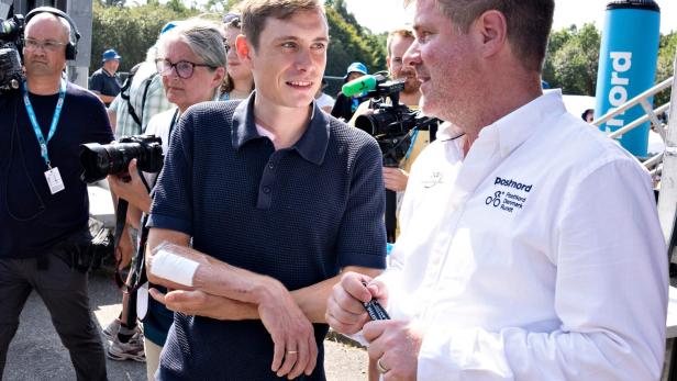 Jonas Vingegaard mit einem Verband am Arm im Gespräch mit einem Mann in einem PostNord-Hemd vor Kameras.