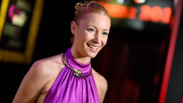 Lachende Frau im violetten Kleid vor unscharfem, bunt beleuchtetem Hintergrund.