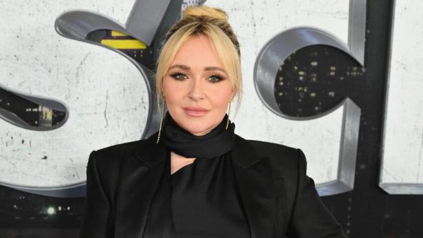 Blonde Frau in schwarzem Outfit mit Mantel und hochgesteckten Haaren vor einer Filmkulisse.