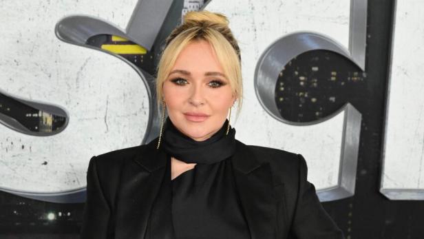 Hayden Panettiere in schwarzem Outfit mit Mantel und hochgesteckten Haaren vor einer Filmkulisse.