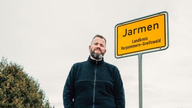 Ein bärtiger Mann steht neben einem gelben Ortseingangsschild der Stadt Jarmen im Landkreis Vorpommern-Greifswald an einer Landstraße.