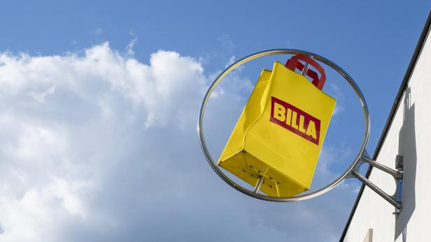 Ein gelbes, taschenförmiges Billa-Firmenschild hängt in einem Metallring vor einem blauen Himmel mit weißen Wolken.