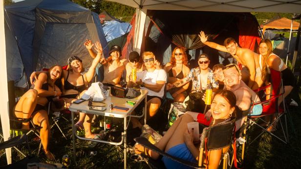 Festivalbesucherinnen und -besucher sitzen lachend im Campingbereich von Frequency zwischen Zelten und Campingstühlen.