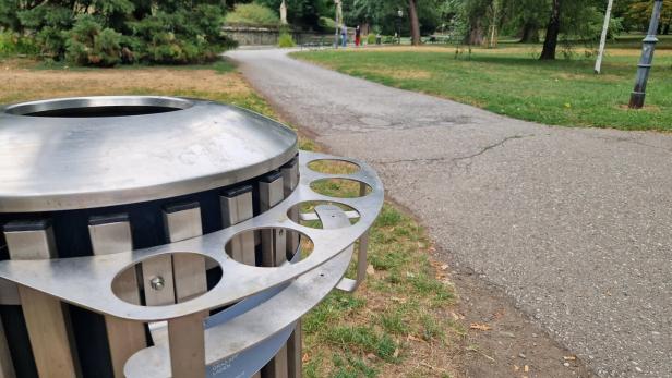 Ein Parkweg führt an einem großen Metall-Mülleimer vorbei, im Hintergrund stehen Bäume und vereinzelte Spaziergänger.