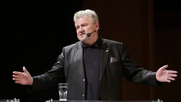 Ein Mann mit Mikrofon gestikuliert vor einem dunklen Hintergrund.