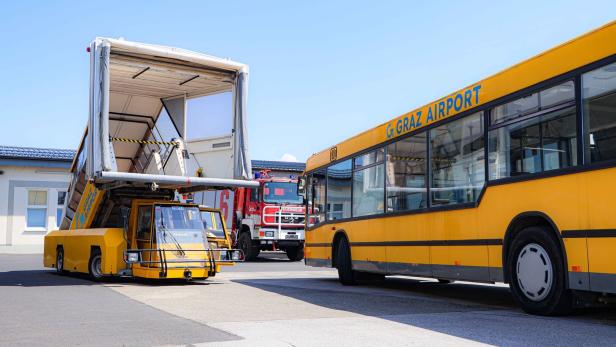 Gelbes Flughafen-Vorfeldfahrzeug und Bus am Graz Airport, dahinter ein Feuerwehrauto.