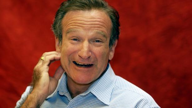 Robin Williams lächelt vor rotem Hintergrund und berührt sich am Ohr.