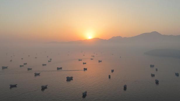 Sonnenaufgang über nebliger See mit vielen Booten und dunkler Bergsilhouette.