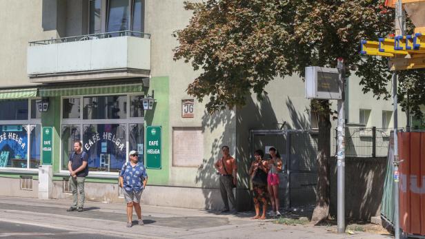 Straßenszene vor dem Café Helga mit mehreren wartenden Passanten, Balkon, Baum und Bushaltestelle.