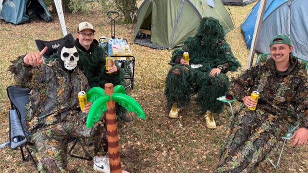 Vier als Waldmenschen verkleidete Frequency-Besucher sitzen mit Bierdosen vor Zelten, neben einer Plastikpalme auf dem Campingplatz.