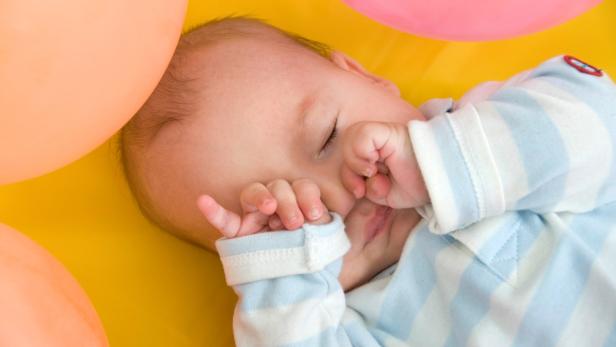 Ein schlafendes Baby liegt auf einer gelben Oberfläche mit bunten Ballons im Hintergrund.
