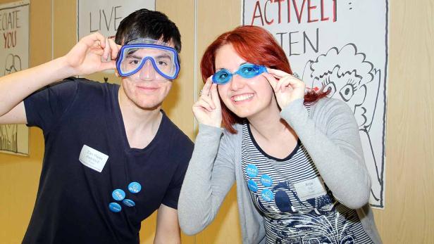 Zwei Personen posieren mit Schwimmbrillen vor einer Wand mit Aufschriften.