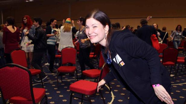 Eine lächelnde Frau steht in einem Raum mit roten Stühlen und vielen Menschen.