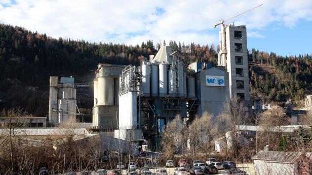 Eine Zementfabrik mit Silos und einem Kran vor einem bewaldeten Hügel.