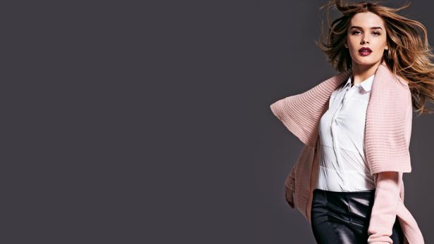 Eine Frau mit wehendem Haar trägt eine weiße Bluse, eine Lederhose und eine rosa Strickjacke.