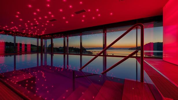 Ein Innenpool mit roter Beleuchtung und Blick auf einen See bei Sonnenuntergang.