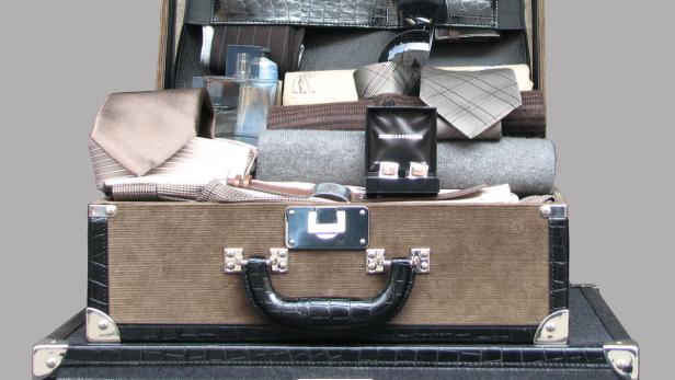 Ein geöffneter Koffer ist gefüllt mit Krawatten, einem Pullover, Manschettenknöpfen und Parfüm.