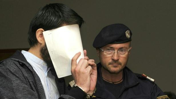 Ein Mann mit Handschellen hält ein Blatt Papier vor sein Gesicht, daneben ein Justizbeamter.