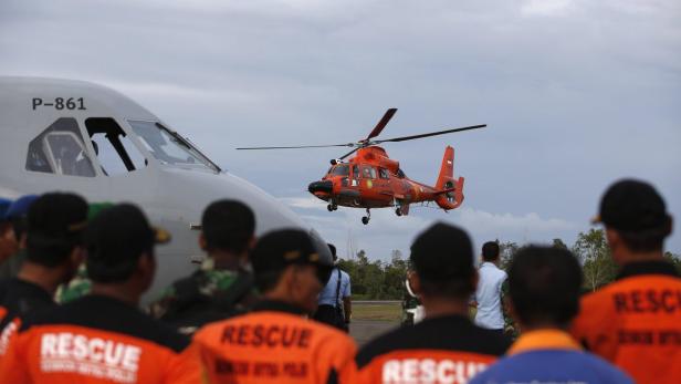 Ein orangefarbener Rettungshubschrauber landet vor einer Gruppe von Rettungskräften und einem Flugzeug.