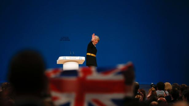 Theresa May winkt von einem Podium vor blauem Hintergrund.