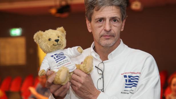 Ein Mann hält einen Teddybären mit einem T-Shirt mit der griechischen Flagge.