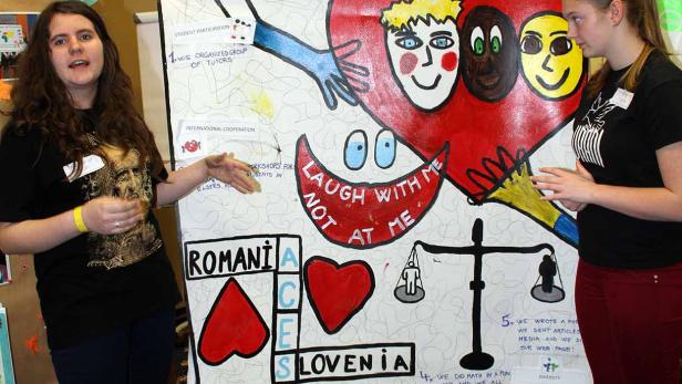 Zwei Frauen stehen vor einem Poster mit der Aufschrift „Laugh with me not at me“.