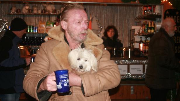 Ein Mann mit Schnauzbart hält einen kleinen, weißen Hund in seiner Jacke und eine Tasse in der Hand.