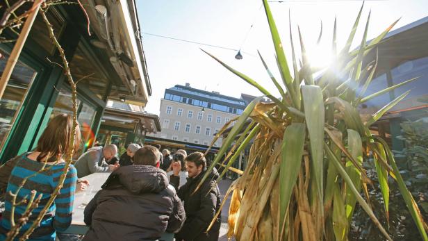 Menschen sitzen an Tischen vor einem Café an einem sonnigen Tag.
