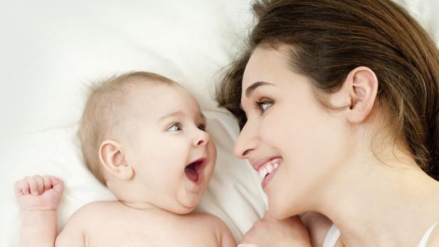 Eine Mutter lächelt ihr lachendes Baby an, während beide auf einem weißen Untergrund liegen.