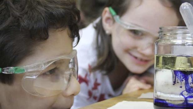 Zwei Kinder mit Schutzbrillen beobachten ein Experiment mit Öl und Tinte in einem Glas.