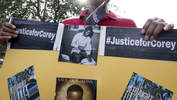 Eine Person hält ein Schild mit der Aufschrift „Justice for Corey“ und weiteren Bildern.