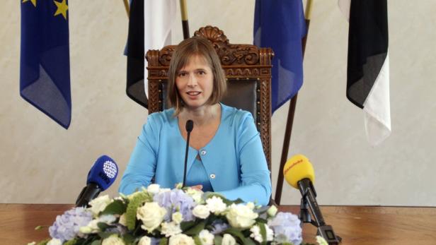 Eine Frau in blauem Blazer sitzt vor Mikrofonen und estnischen Flaggen.