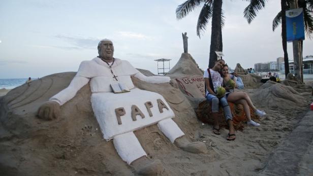 Ein Paar sitzt vor einer Sandskulptur von Papst Johannes Paul II. am Strand.