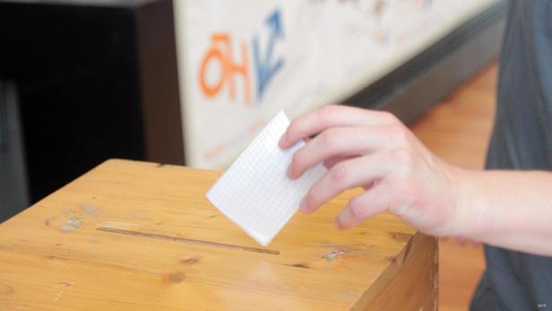 Eine Hand wirft einen Zettel in eine hölzerne Wahlurne.