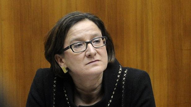 Eine Frau mit Brille und dunkler Jacke vor einem hölzernen Hintergrund.