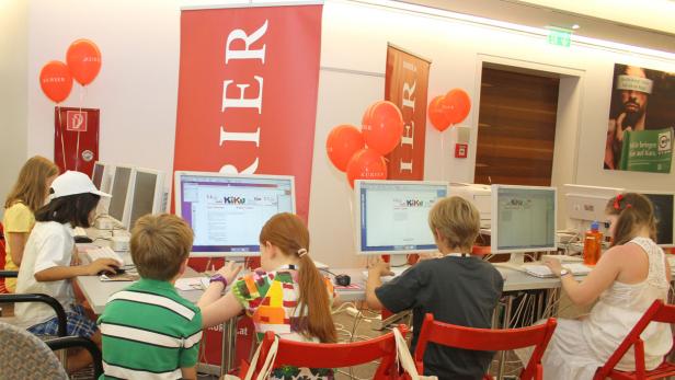 Kinder sitzen an Computern mit dem Logo des „Kurier“ im Hintergrund.