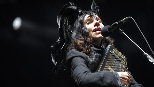 Eine Sängerin mit Federschmuck singt mit einer Zither in der Hand in ein Mikrofon.