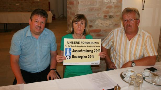 Drei Personen sitzen an einem Tisch und halten ein Schild zur Forderung des Baus der A5 Weinviertelautobahn.