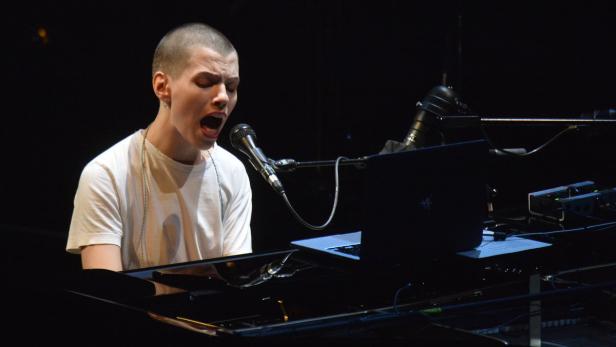 Ein Sänger mit rasiertem Kopf singt am Klavier in ein Mikrofon.