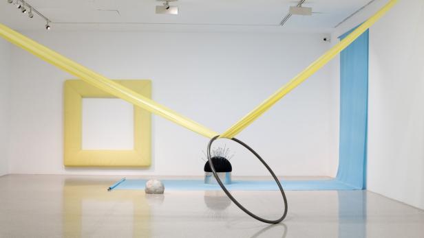 Eine Kunstinstallation mit gelben Stoffbahnen, einem Rahmen und einem schwarzen Ring in einem Ausstellungsraum.