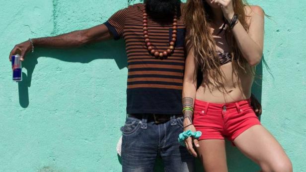 Ein Mann mit Turban und Bart und eine Frau mit roter Hose posieren vor einer türkisfarbenen Wand.