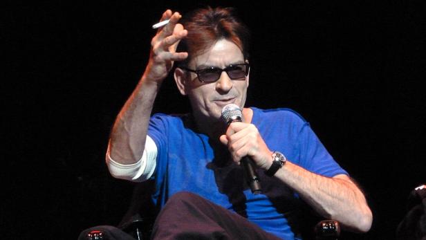 Charlie Sheen mit Sonnenbrille, Mikrofon und Zigarette in der Hand.