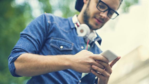 Ein junger Mann mit Brille und Kopfhörern um den Hals schaut auf sein Smartphone.